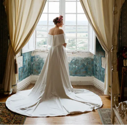La mariée est de dos et regarde par la fenêtre du château et sa traine est étalée sur le parquet. Crédit photo : Alex Schmautz